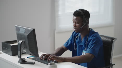 a black man sits at a computer in a doctor's uniform and writes a patient's card while taking calls with headphones. ambulance hotline receive calls and distribute ambulances