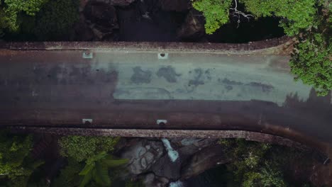 Two-white-vehicles-driving-along-an-old-bridge-in-the-forest-with-waterfall-underneath