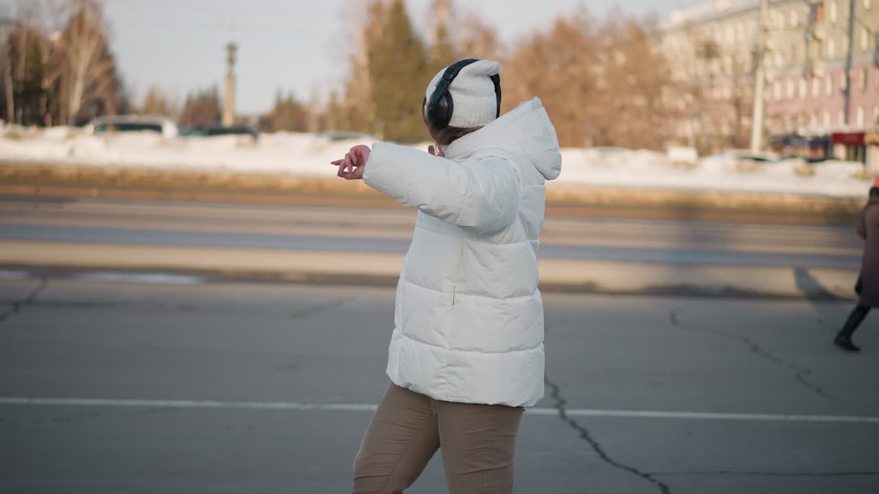 Side view of young artist in white winter coat dancing on cracked pavement, arms spread wide in delight, surrounded by snowy urban scenery, passing vehicles, and pedestrians during sunny winter day