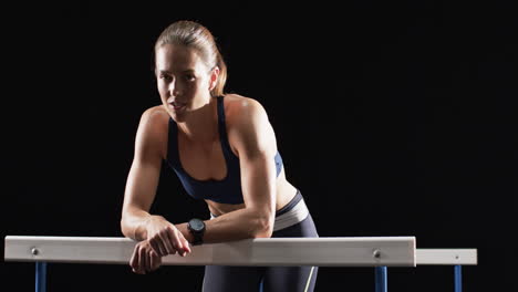 Young-Caucasian-woman-leaning-on-hurdles-during-her-workout-on-a-black-background,-with-copy-space