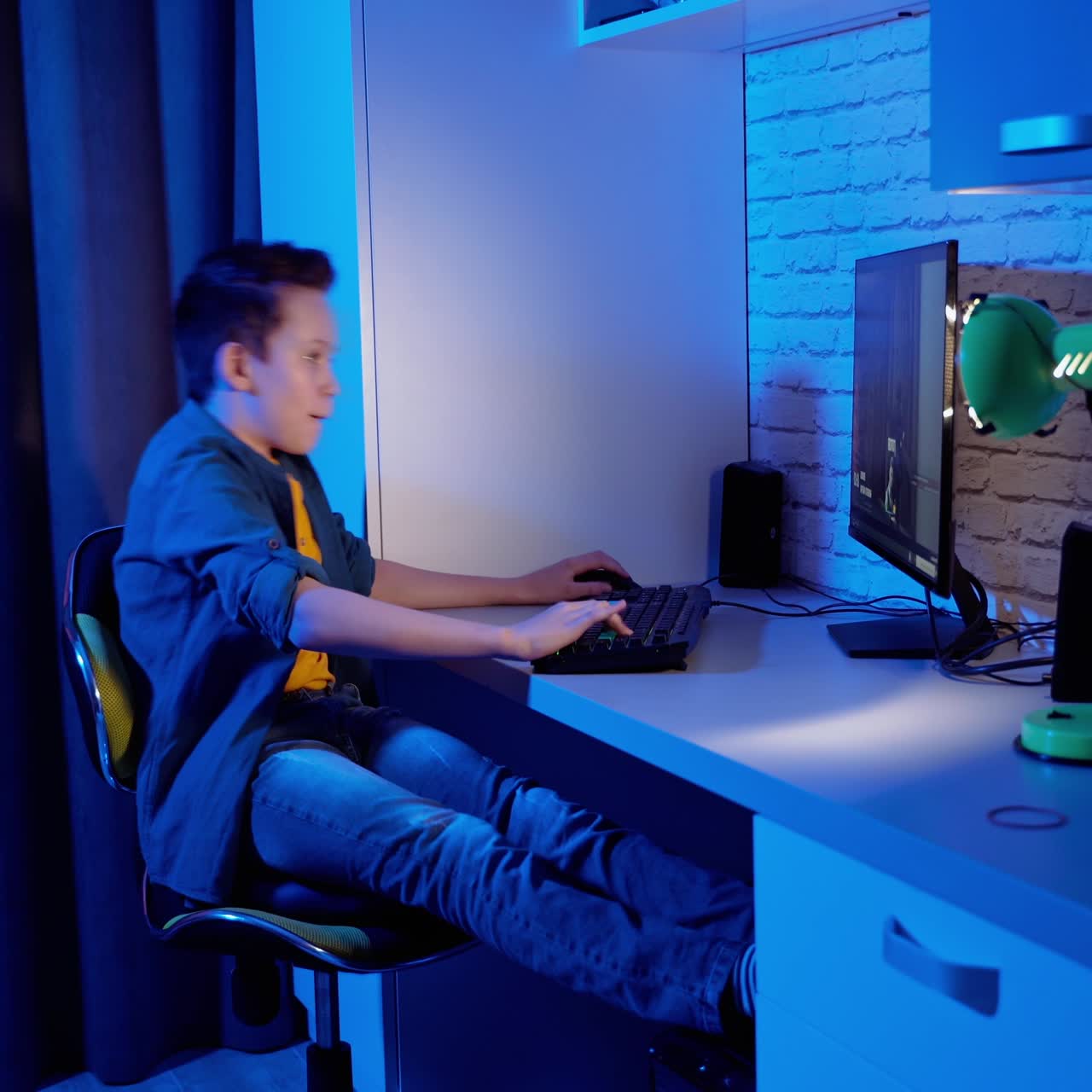 Excited boy playing games on a computer at home. Teen gamer sitting at the table in his room and playing online video games. Blue background.