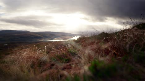 Schnelle-Winde-Zerschlagen-Gras-An-Der-Seite-Eines-Schottischen-Berges,-Während-Lichtstrahlen-Durch-Dunkle-Wolken-Im-Hintergrund-Brechen,-Um-Süßwasserlöcher-Zwischen-Den-Bergen-Hervorzuheben