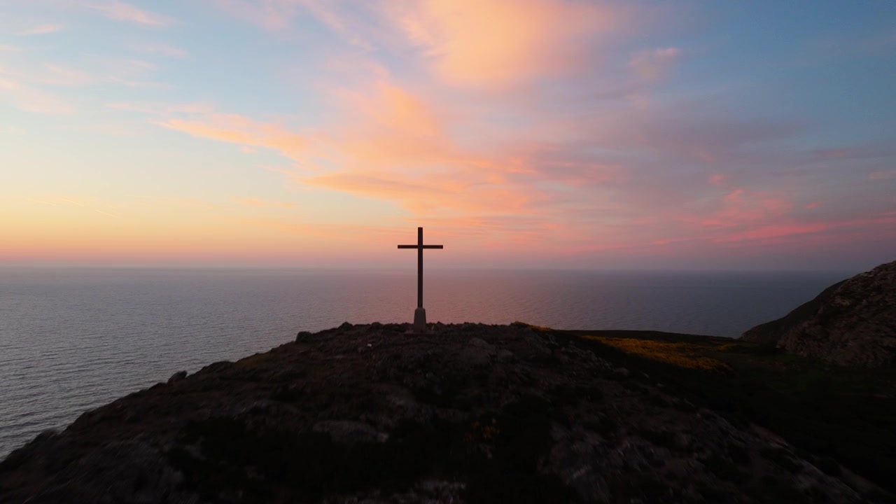 Una cruz solitaria se alza en una colina rocosa mientras el crepúsculo tiñe el cielo de rosa y azul sobre el vasto océano. Ideal para temas de soledad y reflexión