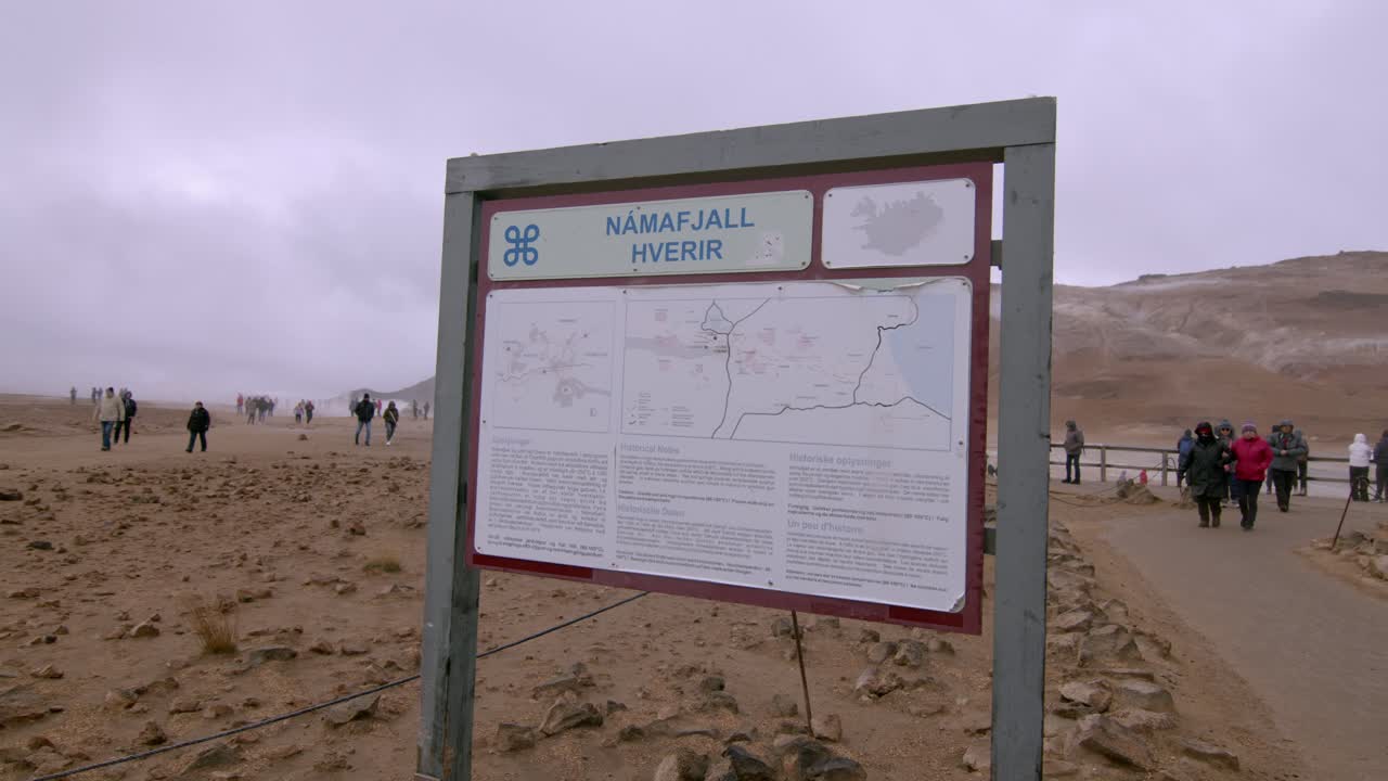 Signboard for Námaskaro hot springs in a barren Icelandic landscape with tourists in the distance