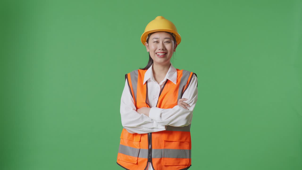 안전 헬을 입은 아시아 여성 엔지니어가 팔을 고 녹색 스크린 배경 스튜디오에 서서 카메라를 향해 미소 짓고 있습니다.