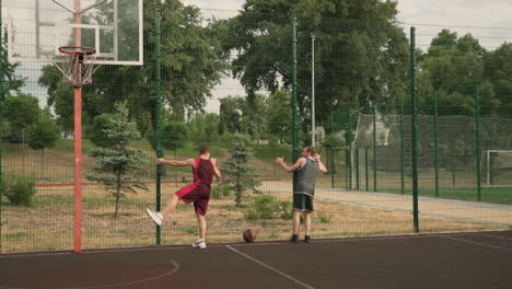 Dos-Jugadores-De-Baloncesto-Masculinos-Estirando-Las-Piernas,-Parándose-Y-Apoyándose-Contra-Una-Cerca-De-Metal-En-Una-Cancha-De-Baloncesto-Al-Aire-Libre