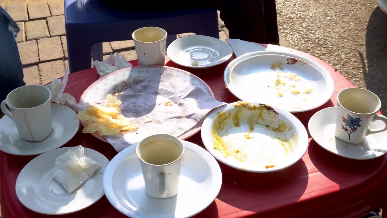 Imagine a bustling local street restaurant where the morning rush has left a breakfast table overflowing with unwashed plates and cups, capturing the essence of busy city life.