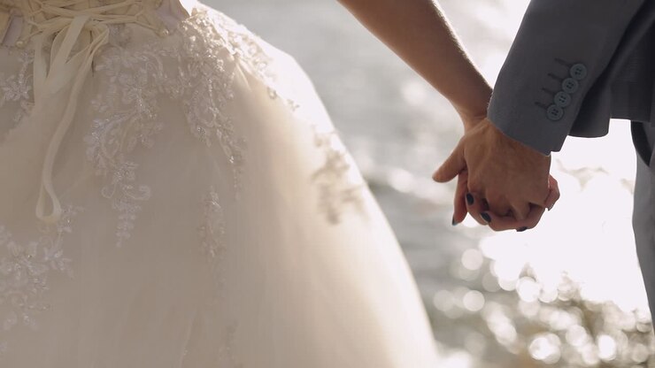 Newlyweds are standing by the sea. They give each other hands. Groom and bride