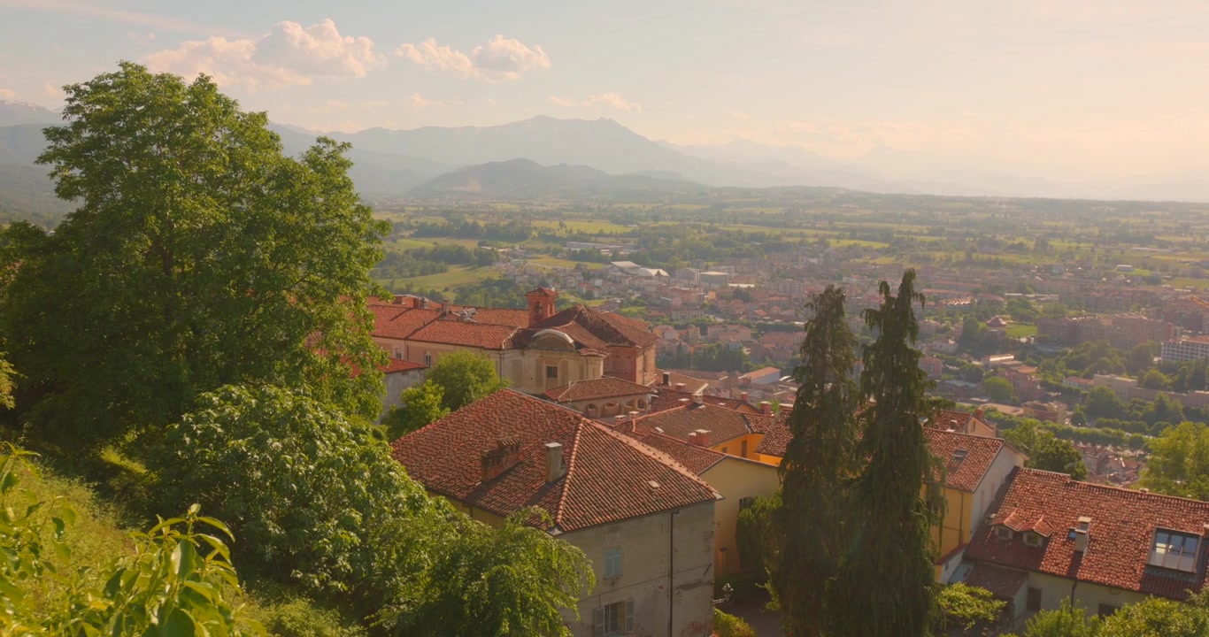 I tetti di tegole rosse di Mondovì si ergono sopra un'ampia valle verde e montagne distanti nella calda luce del tramonto.