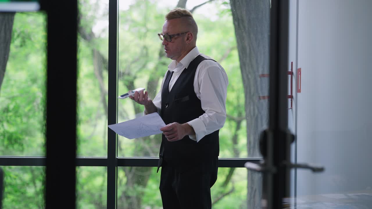 gerente caucasiano profissional gravando correio de voz em smartphone de pé no escritório com documentos. retrato de homem especialista sério falando analisando papelada na janela dentro de casa.