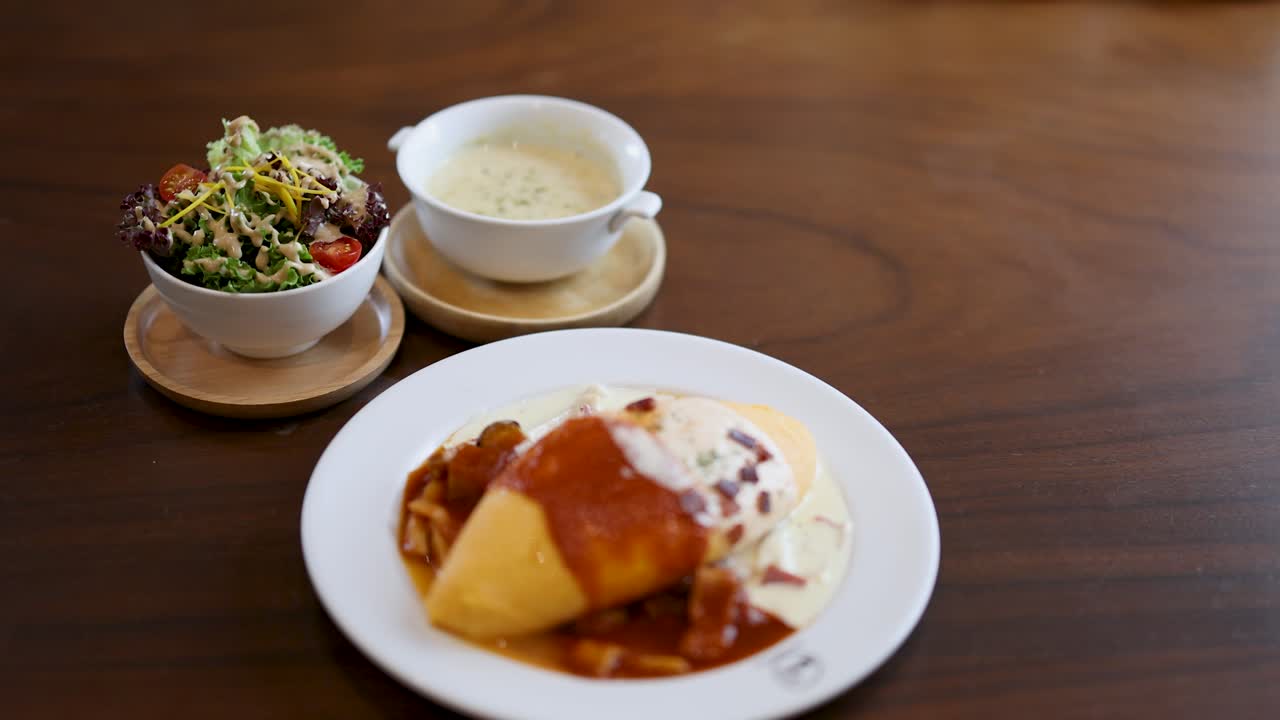 Hand reaches for creamy omelette plate beside salad and soup, natural light, wooden table