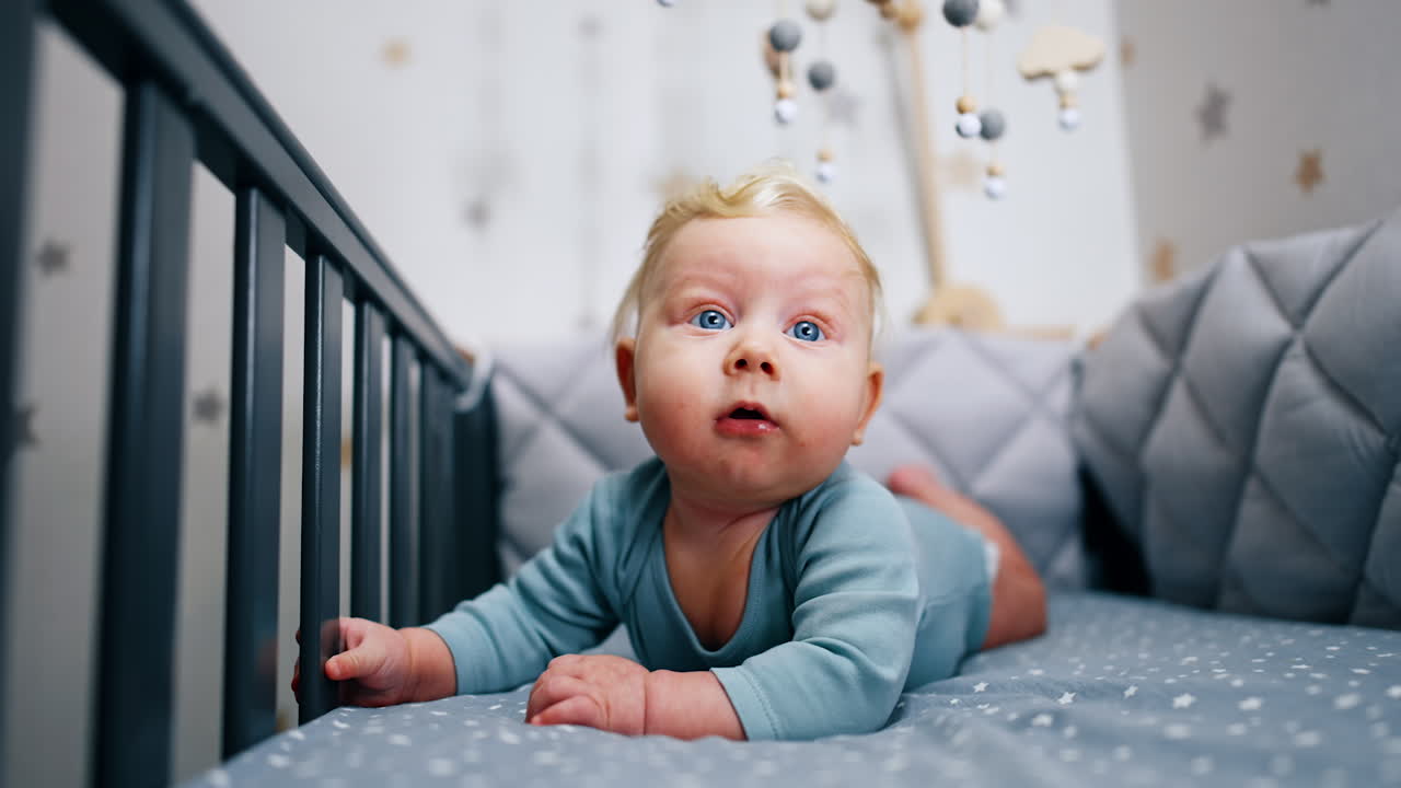 Blond Caucasian infant boy in blue bodysuit lies on the belly in the crib. Adorable blue-eyed boy looking up with surprise.