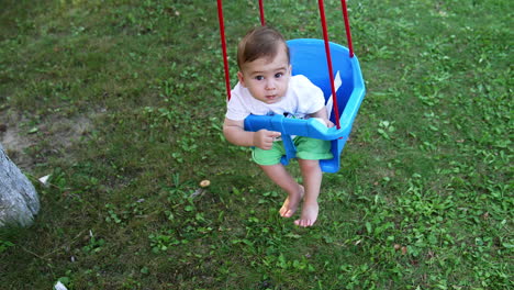sweet little caucasian boy looking into camera swaying in a swing. high angle view. green grass at backdrop.