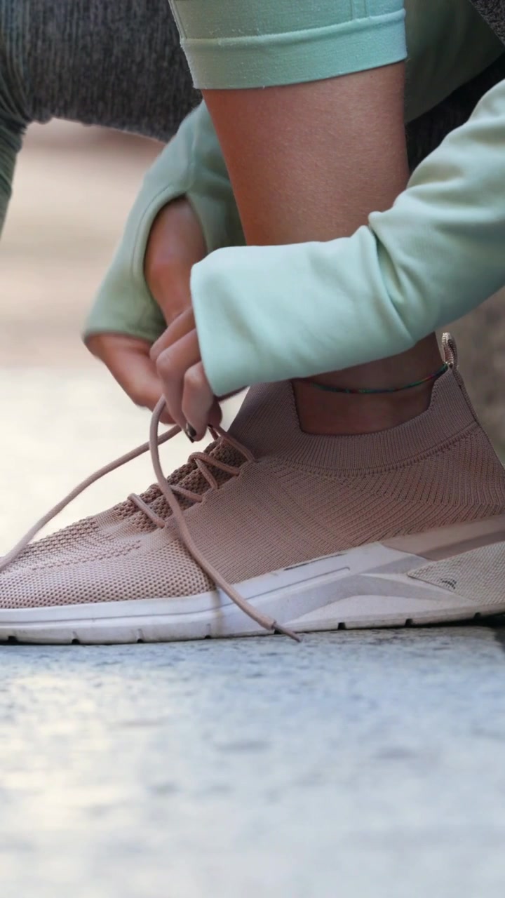 Video verticale: Donna che stringe i lacci delle scarpe annodandoli su un pavimento piastrellato, con un kit color menta preparato prima della corsa.
