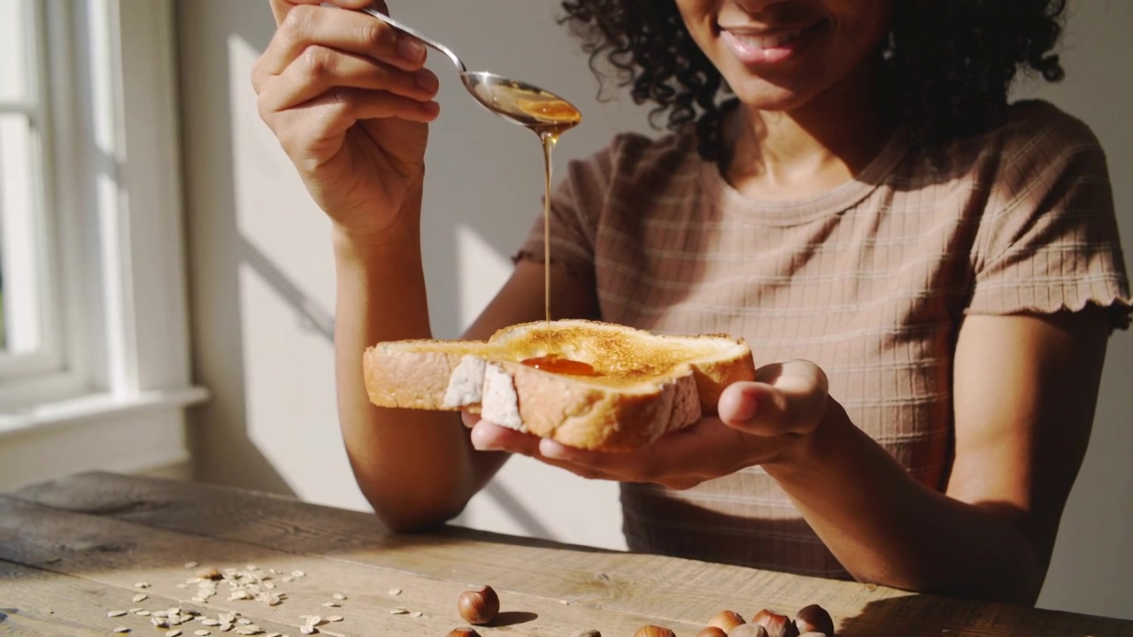 Tilting spoon, woman in ribbed top drizzling honey onto toast at wooden table making breakfast
