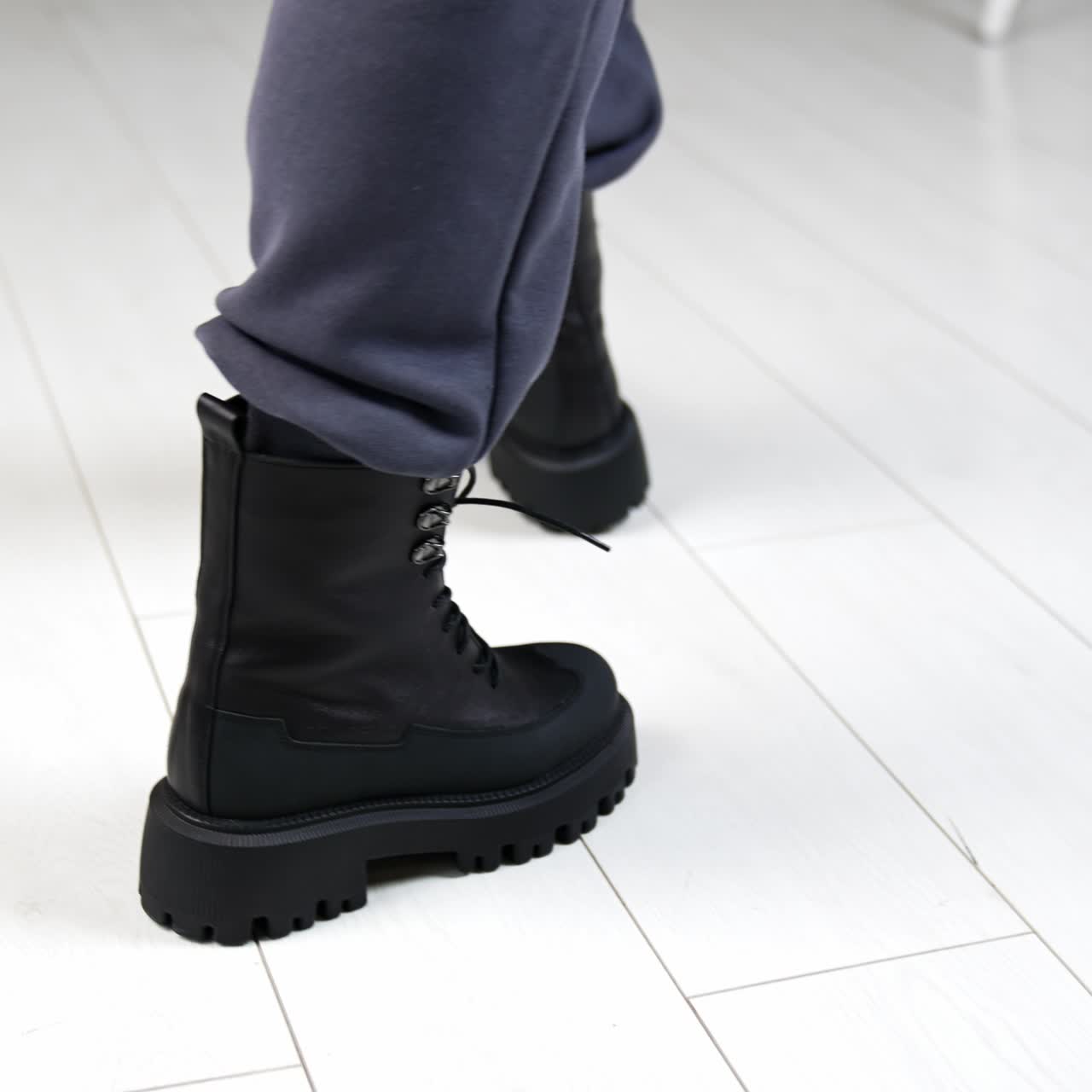 Black massive army boots with laces on model's legs. Woman walks in stylish footwear on the white floor in studio. Top view close up