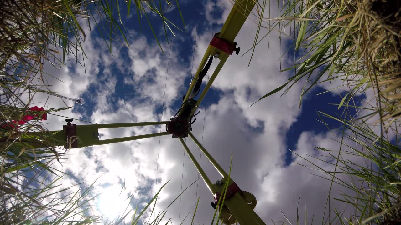 GoPro timelapse from ground looking up at survey tripod with GNSS base station. Clouds moving across blue sky, green grass at frame edges under partly cloudy daylight
