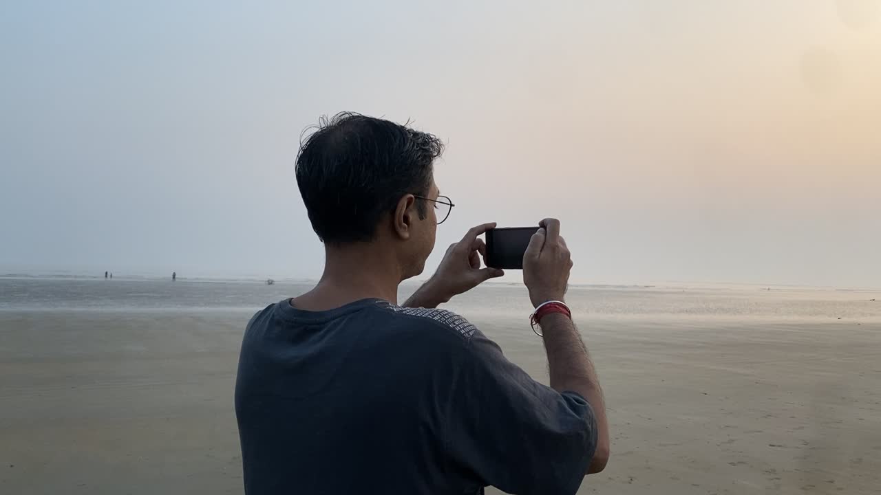 Side view Slow motion gimble shot of an Indian man in blue t-shirt standing in a pleasant beach clicking sunset pictures.