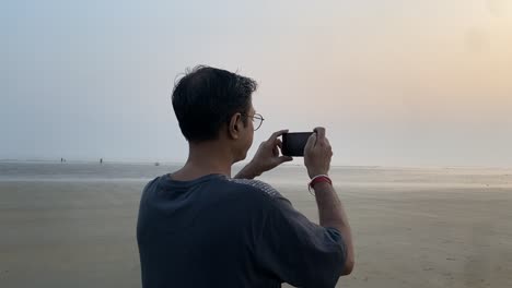 side view slow motion gimble shot of an indian man in blue t-shirt standing in a pleasant beach clicking sunset pictures.