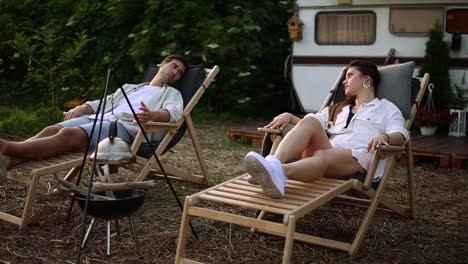 european young couple having rest close to a campsite with hanged kettle . beautiful couple in love laying comfortably on trestle beds and having a talk. rest in nature. modern trailer on background