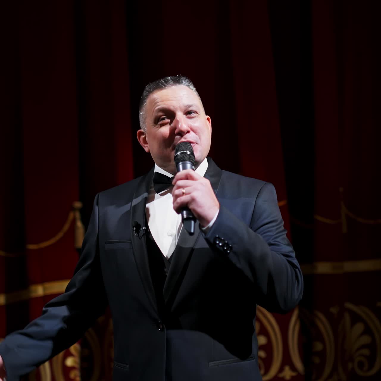 Male announcer with microphone on the stage. Actor in black suit on red scene curtains background