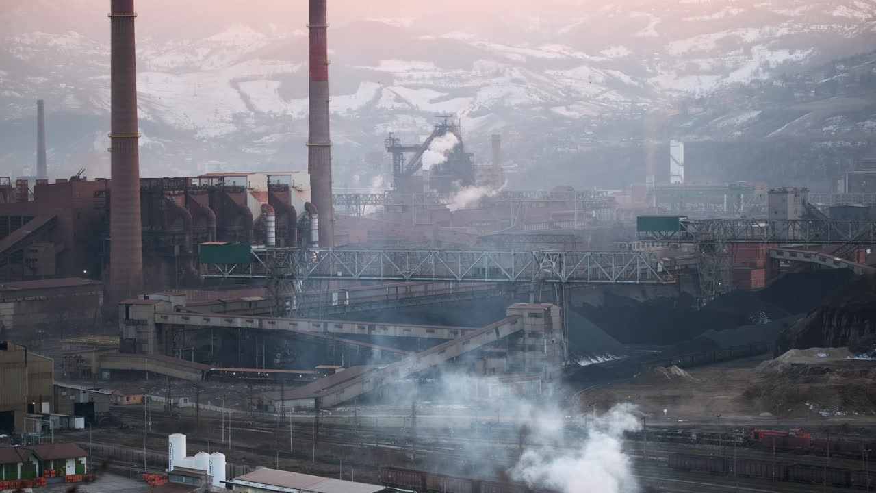 Dawn light illuminates an expansive industrial site with smokestacks and conveyors, framed by a rugged mountainous backdrop