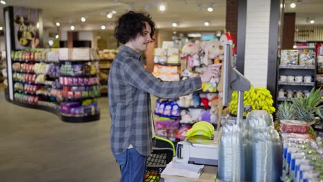 Happy-guy-with-curly-hair-in-a-plaid-shirt-weighs-bananas-using-scales-in-a-supermarket-while-shopping-in-a-grocery-store