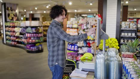 un tipo feliz con el cabello rizado en una camisa a cuadros pesa plátanos usando una balanza en un supermercado mientras hace compras en una tienda de comestibles