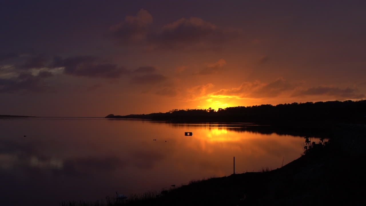 Slow panning shot from left to right of glowing sunset over Malltraeth estuary on Anglesey, North Wales. Vibrant sky colors reflect on calm water creating a scenic coastal travel and nature landscape