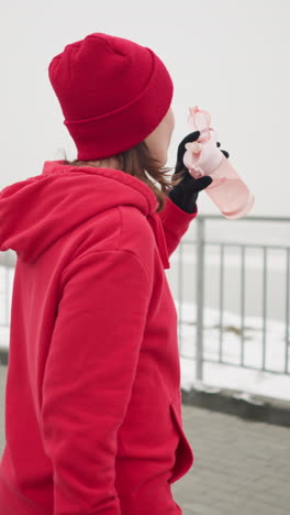 lady in red hoodie and beanie sipping water from pink bottle outdoors during winter walk, gently dropping bottle in serene setting with snow-covered path, iron railing, misty atmosphere in background