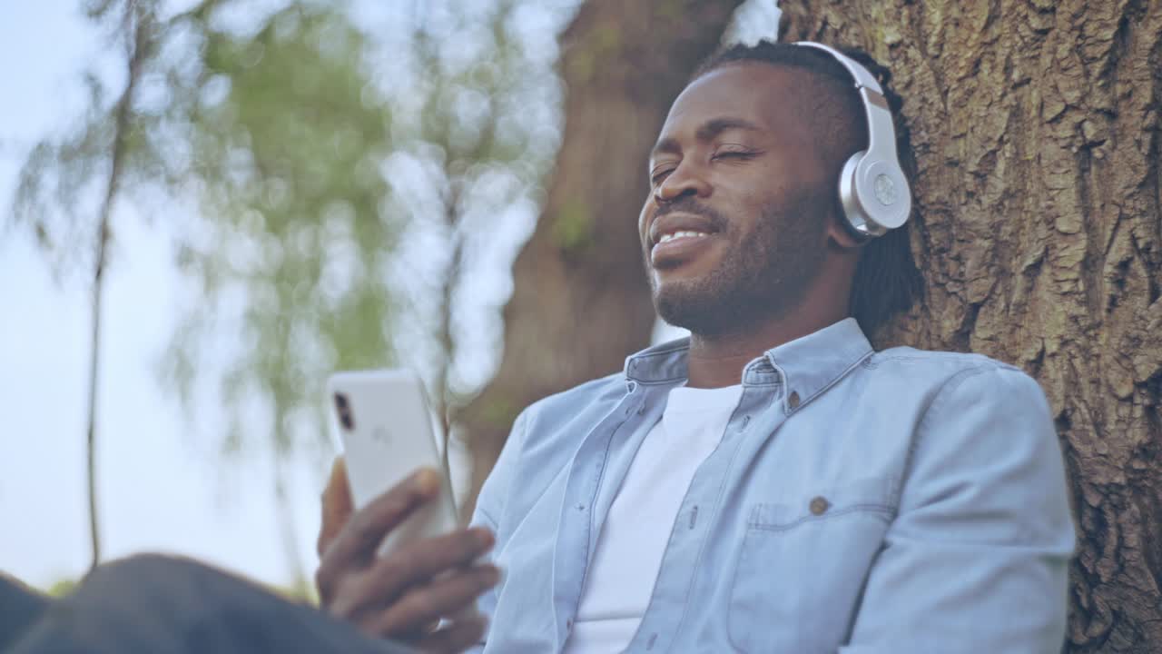 Pleased african-american man in headset listening to music on cellphone in park