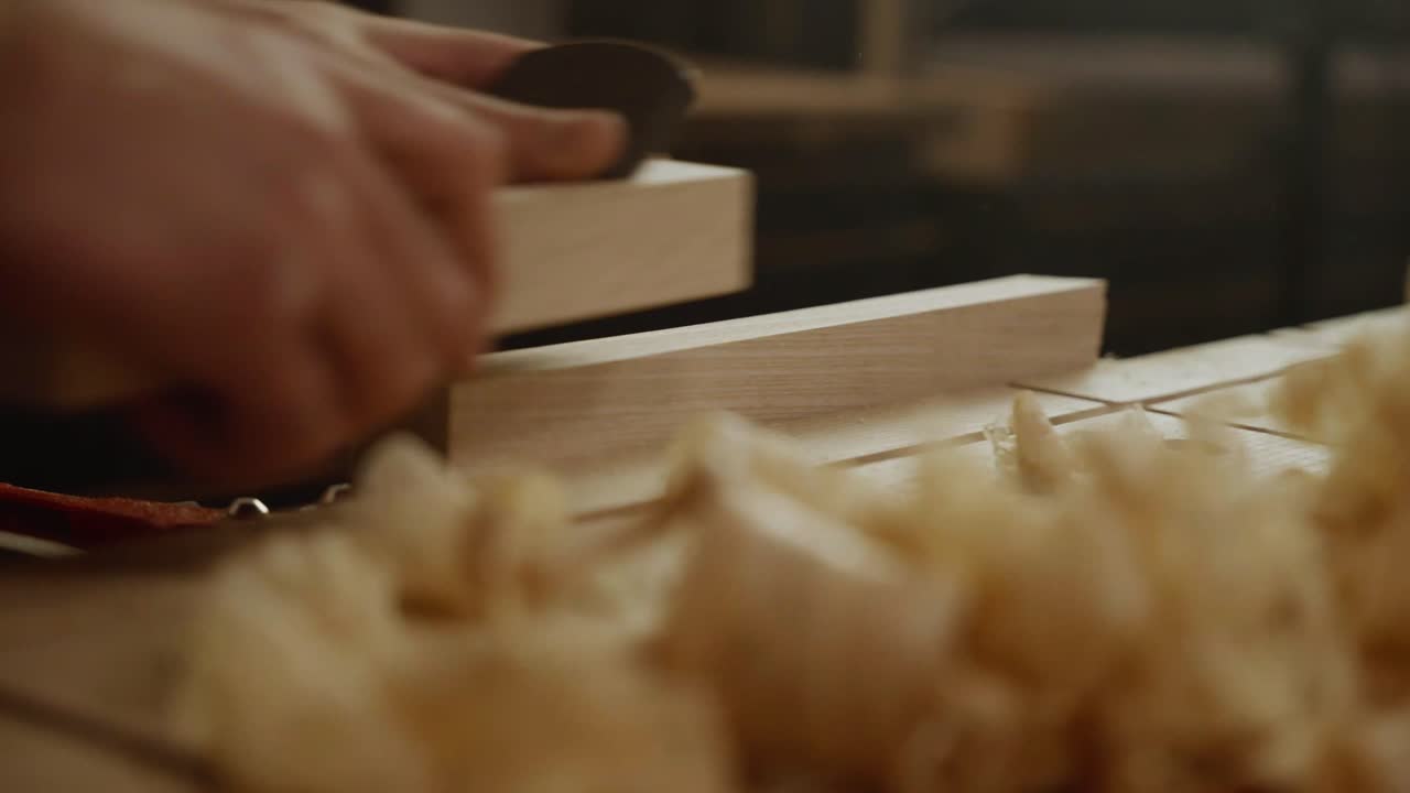 Close-up of a craftsman’s hands planing a wooden board with wood shavings scattered on the workbench, symbolizing craftsmanship, precision, and traditional woodworking