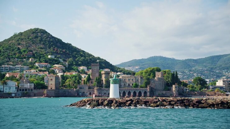 Chateau de la Napoule con le sue torri in pietra e archi sul mare, un faro portuale su un frangiflutti e una collina verde, Mandelieu-la-Napoule, Francia