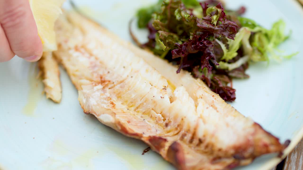 Hand squeezes fresh lemon over grilled fish fillet and mixed greens, shallow depth of field
