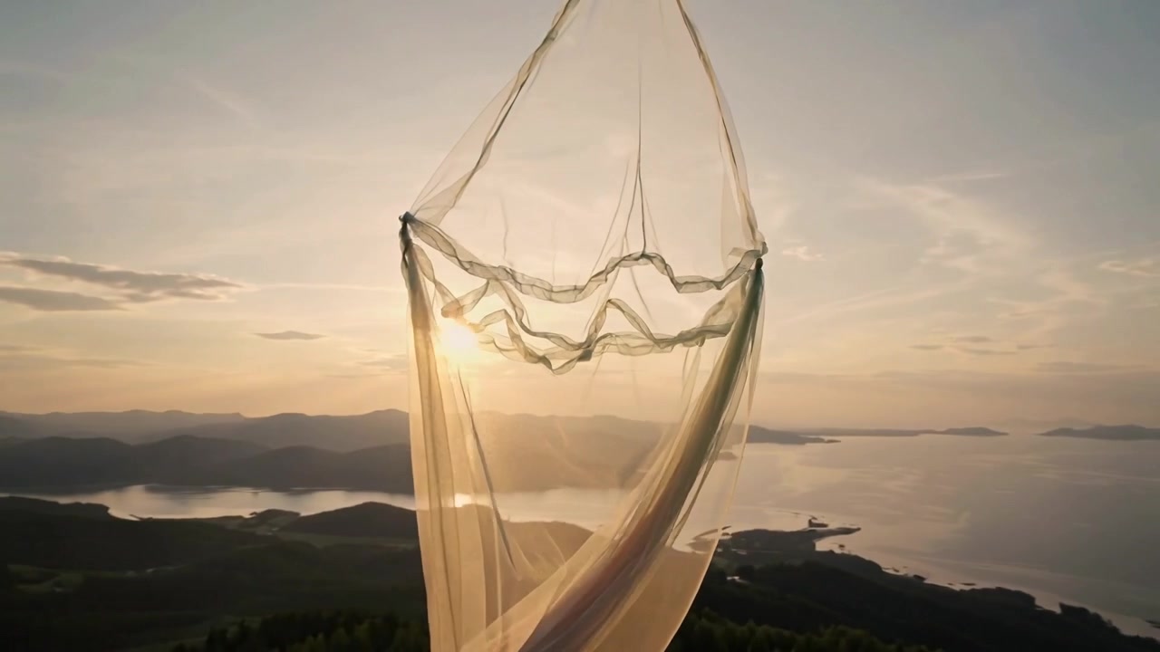 Brezza che solleva un tessuto trasparente sospeso da un anello a goccia sopra una baia costiera, con il sole che retroillumina le isole.