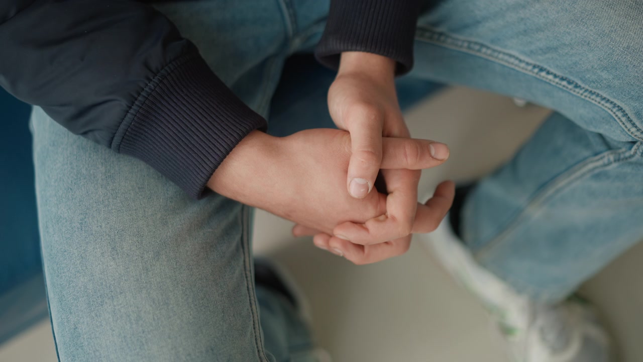 Primer plano de las manos entrelazadas de una mujer joven, vistiendo jeans azul claro y mangas de chaqueta oscura. La escena muestra una iluminación suave con tonos de piel naturales visibles en la textura de las manos.