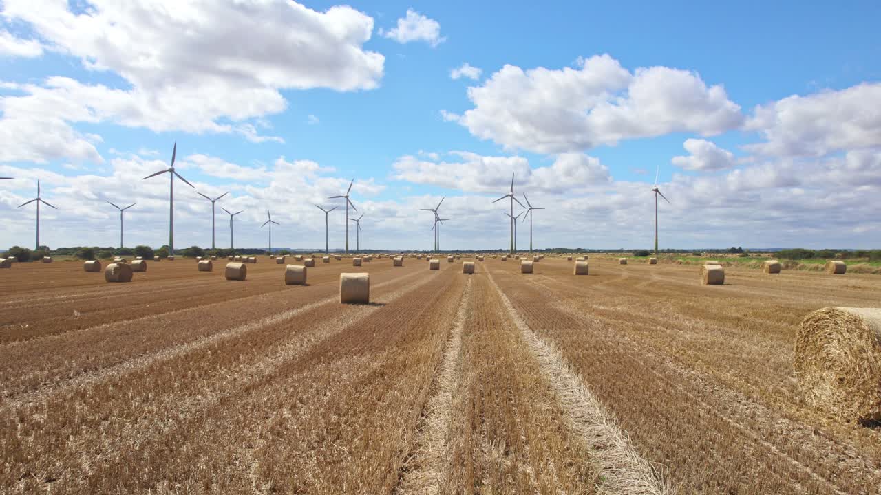 arriba en el cielo, un video aéreo se despliega, mostrando una fila de turbinas eólicas en lincolnshire, girando elegantemente dentro de un campo recién cosechado, adornado con balas de heno doradas