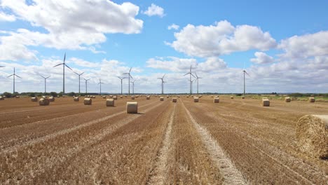 Up-in-the-sky,-an-aerial-video-unfolds,-showing-a-row-of-wind-turbines-in-Lincolnshire,-elegantly-spinning-within-a-field-freshly-harvested,-adorned-with-golden-hay-bales