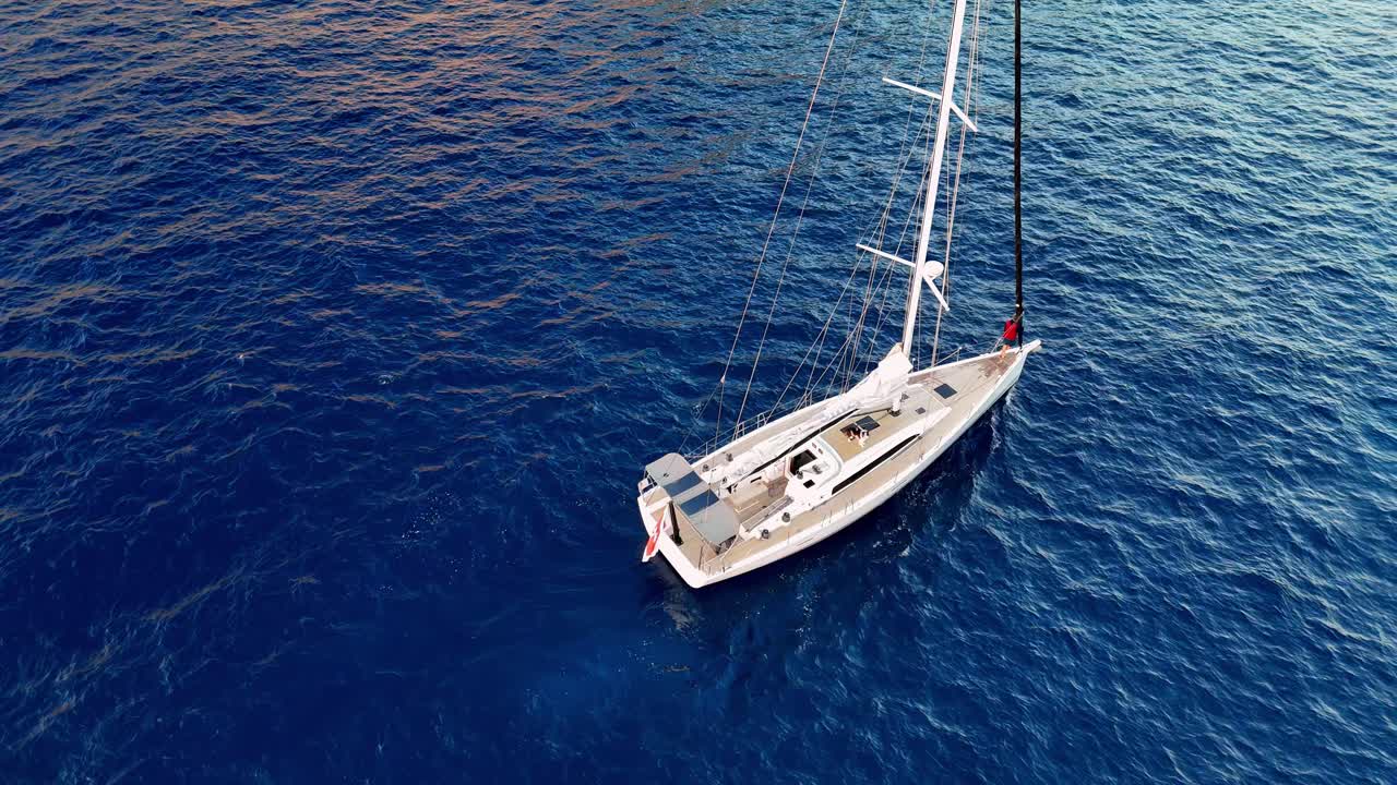 Scatto aereo ad angolazione elevata che cattura un elegante yacht bianco che naviga nelle acque blu intenso del Mediterraneo vicino a Gozo, Malta, durante l'ora d'oro del tramonto estivo, mettendo in mostra vivaci tonalità azzurre e il lussuoso stile di vita nautico