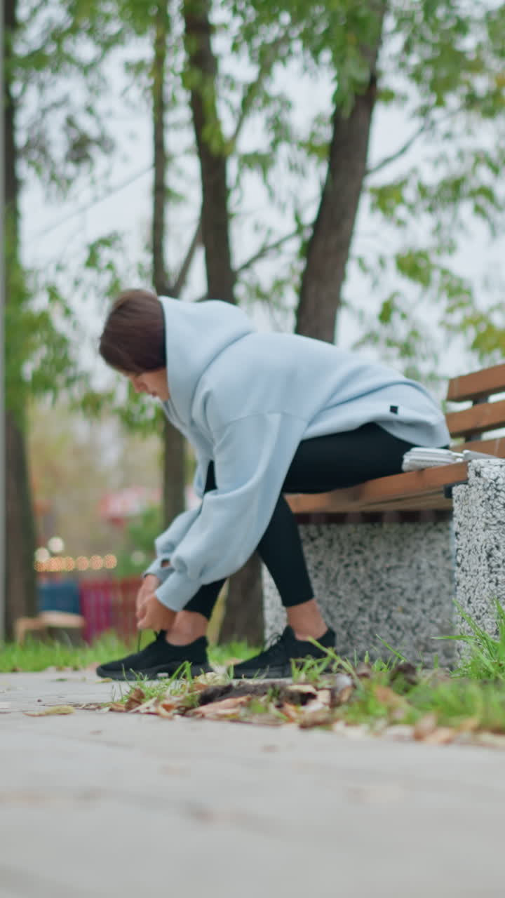 Woman in casual wear sitting on concrete bench tying her shoe, with park background featuring lights, decorations, and trees, creating a serene atmosphere for outdoor activities