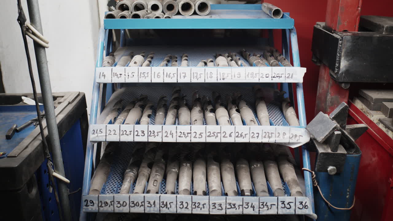 Una rastrelliera per attrezzi da officina con file di grandi punte da trapano industriali in metallo, ordinatamente organizzate ed etichettate per dimensione.