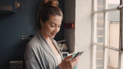 Hermosa-Mujer-En-Pijama-En-Casa-Mirando-Por-La-Ventana-Tomando-Café-Usando-Un-Teléfono-Inteligente