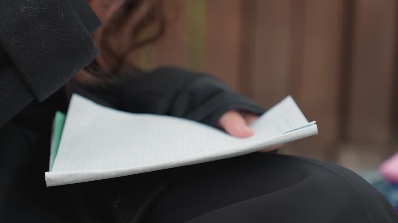 Studerende der skriver i notesbog på bænk udendørs, nærbillede af hænder der holder en pen og vender en side, perlearmbånd der glimter på håndleddet, uldfrakkeærme, skød hvilende på denim, varm træagtig baggrundsbokeh, fokuseret