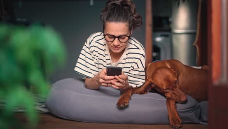 a young woman using her smartphone while lying next to her cute sleeping dog.