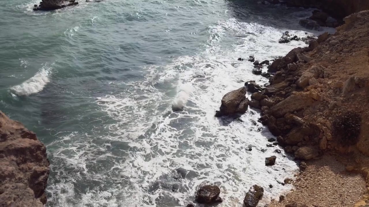 Ripresa aerea con drone di onde marine turchesi che si infrangono contro scogliere rocciose e la costa in una giornata di sole