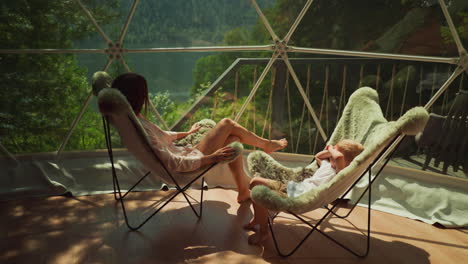 young woman dressed in housecoat rests with toddler son on terrace enjoying breathtaking view. stunning landscape of tranquil lake and dense forest from panoramic window