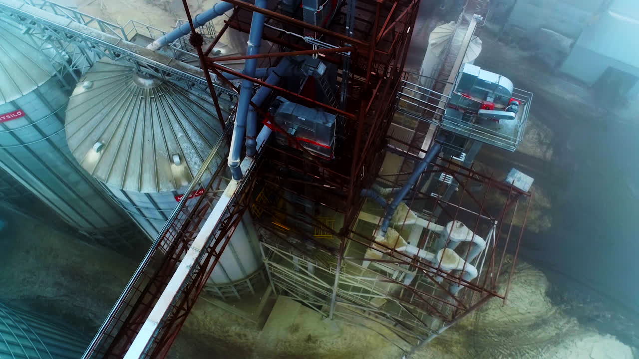 High construction towering over the granary elevator tanks. Flying over the silo complex. Ground covered with snow at backdrop.