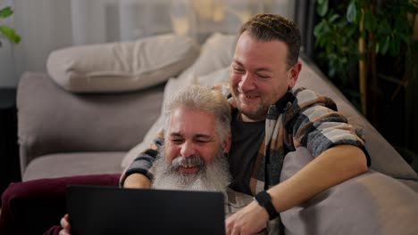 A-happy-elderly-man-with-gray-hair-and-a-lush-beard-lies-in-the-arms-of-his-brunette-boyfriend-with-stubble-in-a-checkered-shirt-and-watches-a-video-using-a-gray-laptop-on-the-sofa-in-a-modern-apartment