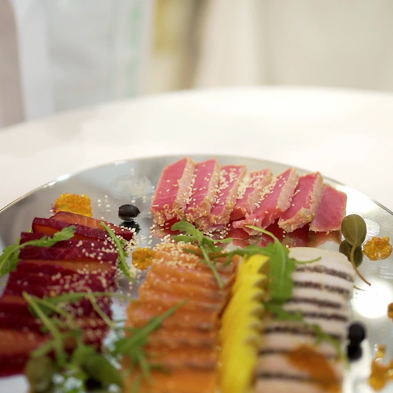 Chef's hand adds cherry tomato to a tasty appetizer of meat on a glass plate on a white table. A cook in white clothes decorates slicing snack for the banquet.