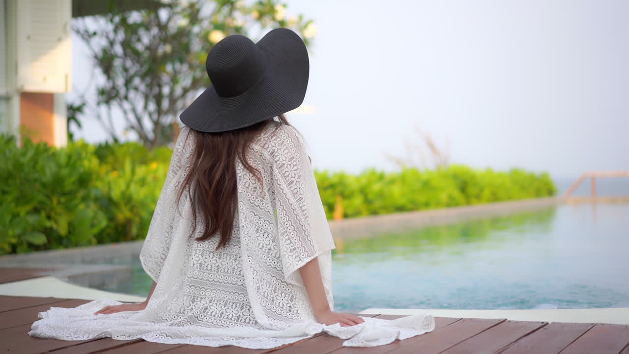 Back of female in summer hat and white cape sitting lonely by the pool on her summer holiday, slow motion